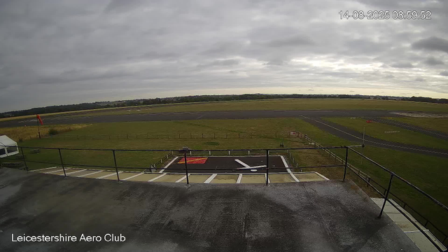 This image shows an outdoor view at the Leicesterhire Aero Club on a cloudy day. In the foreground, there is a parking area with a painted marking on the ground, and a small white tent on the left side. A railing surrounds the edge of a raised platform or deck. Beyond the parking area, there is a grassy field with a red flag and some fencing. Further in the background, a runway or taxiway extends across the scene, with open fields and scattered buildings visible on the horizon. The timestamp indicates the photo was taken on August 14, 2025, at 8:59 AM.