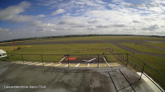 An empty helicopter landing pad with a red marked helipad, surrounded by a railing, is in the foreground. Beyond the pad, there is a grassy area with a windsock on a pole and a small tent or canopy on the left. The background features an open airfield with runways and a partly cloudy sky. The image has a timestamp of "14-08-2025 10:00:13" in the top right corner and "Leicestershire Aero Club" text in the bottom left corner.