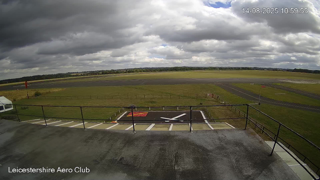 A wide view of an airport runway and surrounding grassy area under a partly cloudy sky. There is a parked helicopter pad marked with an "Y," and the image is labeled "Leicestershire Aero Club" at the bottom. The date and time are displayed in the upper right corner as "14-08-2025 10:59:55."