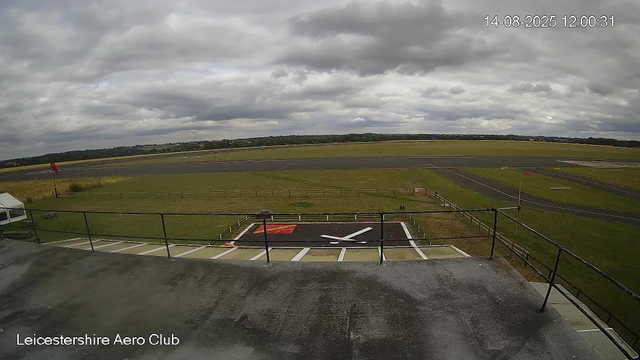 A wide view of an airport tarmac and runway under a cloudy sky. In the foreground, there is a helicopter landing pad marked with a large "H" and red markings. To the left, there is a small white tent, and a windsock on a pole is visible. The area is enclosed by a metal railing. The background features open grassy fields and airport runways stretching into the distance. The timestamp shows the date as August 14, 2025, at 12:00:31 PM, and the label indicates this is from the Leicestershire Aero Club.