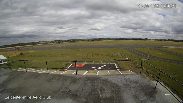 An outdoor view from a webcam at Leicesterhire Aero Club shows a cloudy sky with patches of blue, a runway stretching across the middle distance, a grassy area with a windsock on the left, and a paved helipad marked with a large "H" and a white arrow in the foreground. The timestamp indicates it was taken on August 14, 2025, at 1:00 PM. The overall scene appears calm with no aircraft or people visible.