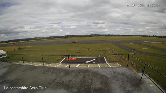 A small aircraft with a yellow tail and blue fuselage is taxiing on the runway at Leicesterhire Aero Club. The sky is partly cloudy, and the surrounding landscape is flat with grassy fields. There is a helicopter landing pad in the foreground, with a prominent white and red marking. The image is timestamped "14-08-2025 14:00:32" in the top right corner.