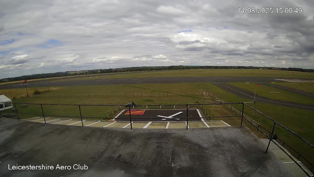 A view from a webcam at Leicesterhire Aero Club shows an overcast sky with thick clouds. In the foreground, there is a helipad with a white helicopter symbol painted on it, surrounded by a black border and enclosed by a railing. To the left, there is a small white tent, and a picnic table is visible nearby. The tarmac of the runway extends beyond, with various markings and signs visible. A wind sock stands on the left side of the image, and the scene is framed by grassy fields and a distant tree line under a cloudy sky. The timestamp in the top right corner reads "14-08-2025 15:00:49".
