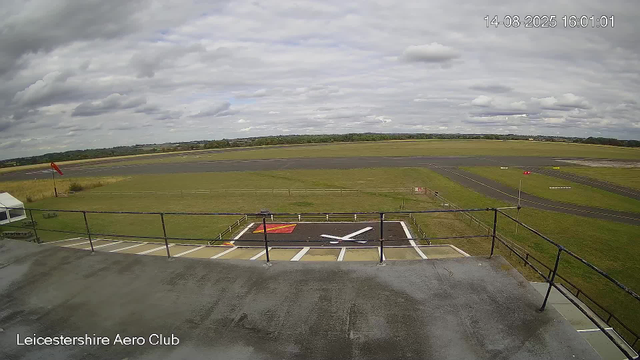A view of an airfield from a webcam, showing a runway in the distance under a cloudy sky with patches of sunlight. In the foreground, there is a helipad with a large white "Y" and a red square. Surrounding the helipad is a grassy area enclosed by a fence, and there is a windsock on a pole to the left. The timestamp in the top right corner reads "14-08-2025 16:01:01," and the bottom left corner has the text "Leicestershire Aero Club."