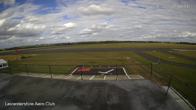 An outdoor view of an airfield with a runway extending into the distance, green fields, scattered clouds in the sky, and a fence in the foreground. There is a helipad marked with a large "H" and a red square, located near the bottom center. The date and time stamp in the upper right corner reads "14-08-2025 17:00:51," and the text "Leicestershire Aero Club" appears at the bottom left corner.