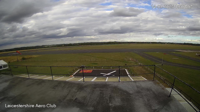 An outdoor view of the runway at Leicestershire Aero Club on a cloudy day. The sky is mostly covered with dark and light clouds, with some patches of blue. The runway extends into the distance, bordered by fields and trees. In the foreground, there is a small helipad with a large white "H" on a black surface, surrounded by a white border and a fence. To the left, there is a small white tent, and to the right, a small aircraft is parked on the grass. The timestamp in the upper right corner indicates the date and time as August 14, 2025, at 6:01 PM.