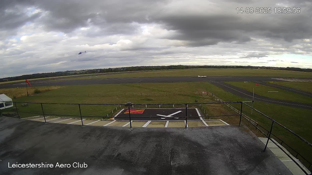 A view from a webcam at Leicesterhire Aero Club showing part of the tarmac runway area, a helipad with a large aircraft propeller symbol painted on it, a grassy field, a small aircraft parked on the grass, and a helicopter flying in the cloudy sky. The scene appears to be late in the day, with overcast weather.