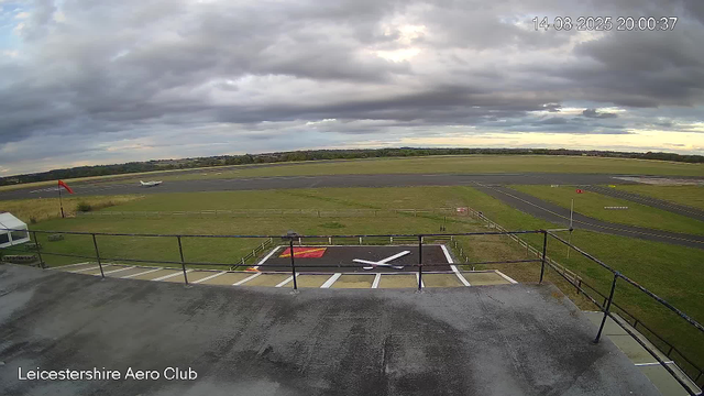 A wide view of an airfield with a small aircraft on the runway, a windsock on the left, and a helicopter landing pad in the foreground. The sky is overcast with dark clouds, and the landscape in the distance is flat with some trees. The timestamp shows "14-08-2025 20:00:37," and "Leicestershire Aero Club" is written at the bottom left corner.