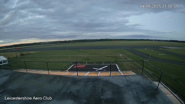 An empty helipad with a large white clock symbol is seen from a webcam view, with a grassy airfield extending into the distance under a cloudy sky. The clock symbol is painted on the helipad, and there is a small red flag on a flagpole nearby. A white tent-like structure is visible on the left side, and a smokestack-like feature is also present. The fence surrounds the helipad area, and the landscape stretches out with open fields and runways. The time and date are displayed in the top right corner: 14-08-2025 21:00:51. The bottom left corner reads "Leicestershire Aero Club."