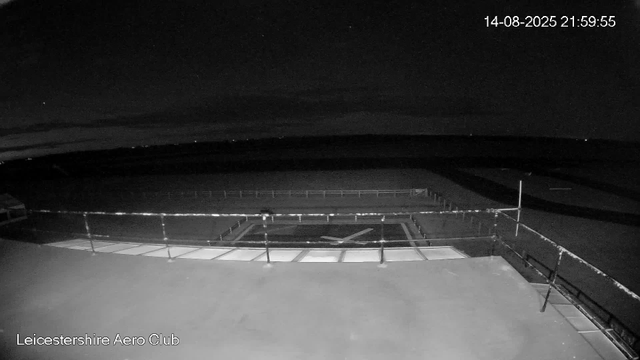 A black-and-white webcam image shows an empty observation deck or balcony at Leicestershire Aero Club during nighttime. The deck has a railing along the edge and a ground surface that appears smooth. The sky is dark, with a few distant stars visible. The timestamp on the top right reads "14-08-2025 21:59:55."