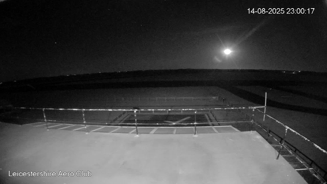 A black-and-white image taken at night shows the rooftop of LeicesterShire Aero Club. The sky is dark with a bright, full moon and a few stars visible. There is a railing around the rooftop's edge, and the scene appears quiet and empty. The timestamp in the top right corner reads "14-08-2025 23:00:17".