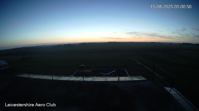 A wide view of an open field at dawn, with the sky gradually brightening and a few scattered clouds. In the foreground, part of a building's rooftop with railings, a helicopter pad marked with an "X," and a small vehicle parked on the left side. The timestamp in the upper right corner shows the date as August 15, 2025, at 5:00 AM. The text at the bottom left corner reads "Leicestershire Aero Club."
