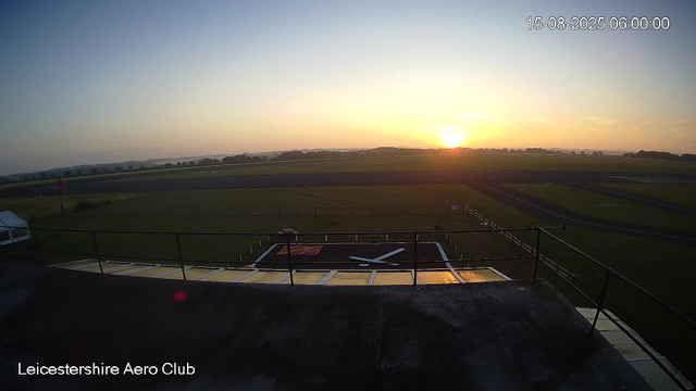 The image shows a view of the Leicestershire Aero Club's airfield at sunrise. The sky is clear with the sun just beginning to rise on the horizon, casting a warm glow over the landscape. In the foreground, there is a small helipad with a large white "H" painted on it, surrounded by a railing. To the left, part of a white tent or shelter is visible. The airfield features grassy areas and a runway extending into the distance, with a few signs and markings along the way. The overall scene is calm and early in the morning.