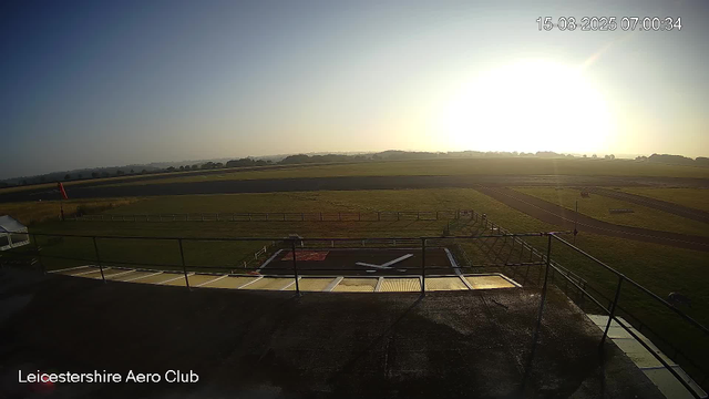 An outdoor view of an airfield taken in the early morning at the Leicestershire Aero Club. The sky is clear with the sun low on the horizon, casting a bright light across the grassy airfield and runway areas. The foreground includes a helicopter landing pad with a large white "H" and surrounding fencing. There are no people visible in the image.