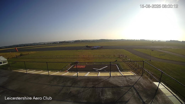 A small airplane is taxiing on a runway at Leicester Aero Club during what appears to be early morning or late afternoon, with the sun low in the sky. The scene is clear, with a grassy field and open sky in the background, and a windsock on the left side of the image. The date and time shown are 15-08-2025 at 08:00:12. The foreground includes a railing and a marked helipad on the ground.