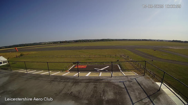 An empty tarmac area with a helipad marked by a white "H" and a red square, surrounded by a black metal fence. Beyond the fence, there is an open grassy field and a runway in the distance. The sky is clear and blue, and the timestamp shows the date as August 15, 2025, at 10:00 am. The location noted is Leicestershire Aero Club.