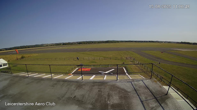 A view from a webcam at Leicestershire Aero Club shows a clear sky with the airfield's runway in the background. In the foreground, there is a rooftop area with a railing, a marked helipad, and a small tent on the left. A person is walking along a fence near the grass, and a windsock is positioned to the left of the image. The scene is calm, with no aircraft visible. The timestamp indicates the date and time as August 15, 2025, at 11:00:48 AM.
