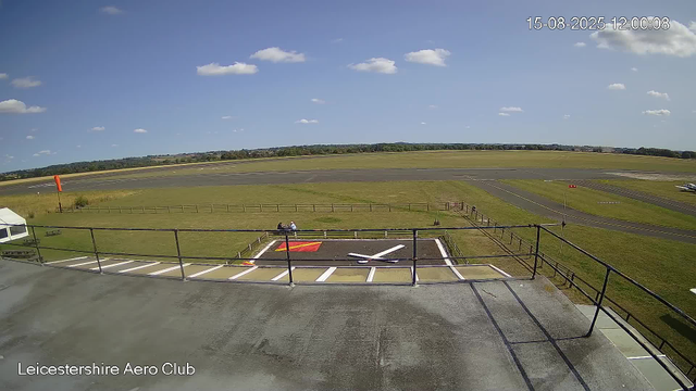 A clear day at Leicesterhire Aero Club with blue skies and scattered white clouds. The airfield is visible with a runway extending into the distance, surrounded by grassy areas. In the foreground, there is a small fenced area with a white and red helicopter rotor blade lying on the ground, and a person sitting on a bench nearby. To the left, a tent is set up, and a windsock is visible on a pole. The scene is peaceful and open, with a few small aircraft parked on the right side of the airfield. The timestamp shows the date and time as August 15, 2025, at 12:00:08.