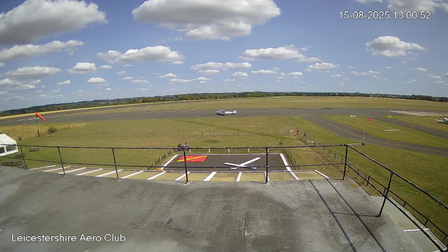 A view from a camera at Leicesterhire Aero Club showing a runway with a small aircraft taxiing on it under a partly cloudy sky. In the foreground, there is a parking area with marked spaces, a small white tent, a bench, a red and yellow windsock, and a sign with parking instructions. The horizon is lined with trees and open fields. The timestamp indicates the photo was taken on August 15, 2025, at 1:00 PM.