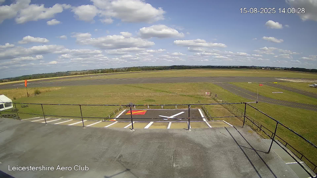 An outdoor view of an airfield under a partly cloudy sky, with a large grassy area and a runway in the background. In the foreground, there is a viewing platform with safety railings, a designated helicopter landing area marked with a bold “H,” and a small white tent. The image is timestamped "15-08-2025 14:00:28" and labeled "Leicestershire Aero Club" in the bottom left corner.
