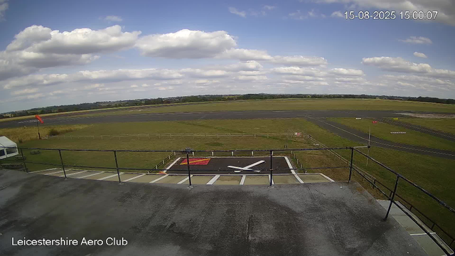 A view from a webcam at Leicesterhire Aero Club showing a runway and surrounding grassy fields under a partly cloudy sky. The date and time are in the top right corner: 15-08-2025, 15:00:07. A helicopter landing pad marked with a white "H" and a red and white wind sock are visible in the foreground, along with some fencing and a small tent.