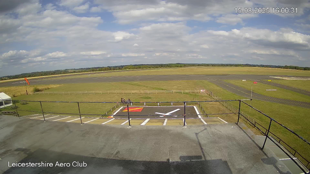 An empty tarmac at Leicesterhire Aero Club under a partly cloudy sky, with a windsock on the left side, a small fenced grassy area with benches in the foreground, and a runway stretching into the distance. The date and time are displayed in the top right corner as 15-08-2025 16:00:31.