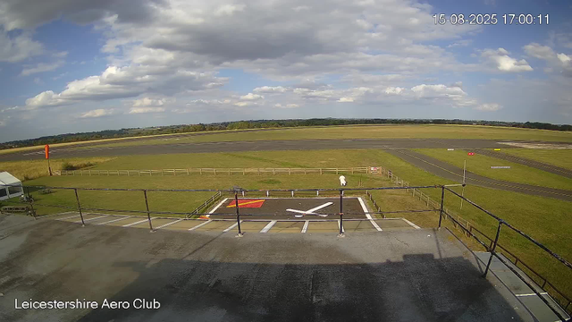 A view from a webcam shows an airfield under a partly cloudy sky. In the foreground, there is a ramp with a small helipad marked with a white helicopter symbol and a red square near the bottom left. A person in white clothing is walking on the grass beyond the ramp, facing away from the camera. The airfield has a runway extending into the distance, surrounded by grassy areas and a distant tree line. The timestamp reads "15-08-2025 17:00:11," and the bottom left corner displays the text "Leicestershire Aero Club."
