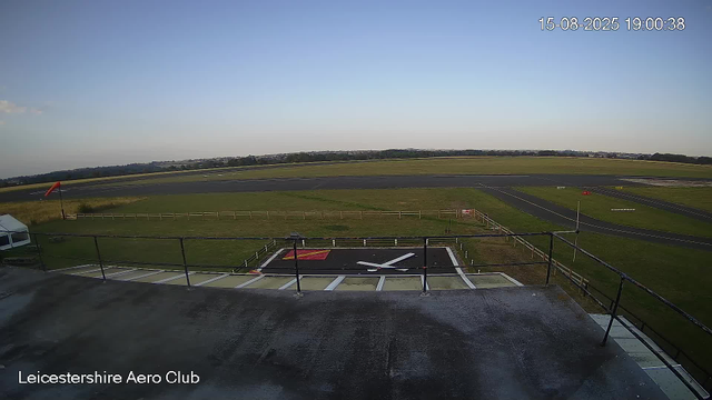 A view from a webcam at Leicestershire Aero Club shows a clear sky over the airport runway and grassy areas. In the foreground, there is a mounted helicopter rotor head with blades and a red and white windsock on a pole to the left. The date and time are displayed in the top right corner as 15-08-2025 19:00:38. The image captures the runway, surrounding grass, and distant horizon with trees and buildings.