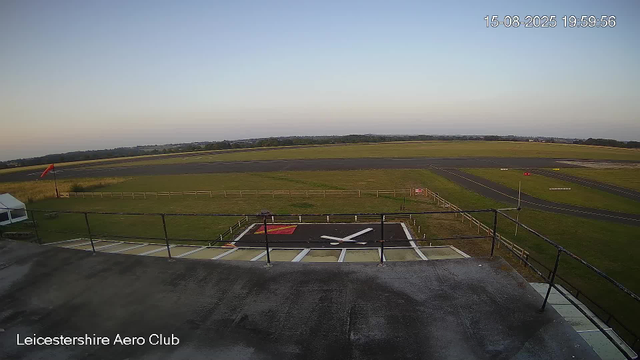 An empty aircraft carrier parking area with a black helipad featuring a white cross and a red square in one corner, surrounded by a fenced green field, with a windsock on a pole and a distant runway under a clear sky at dusk.