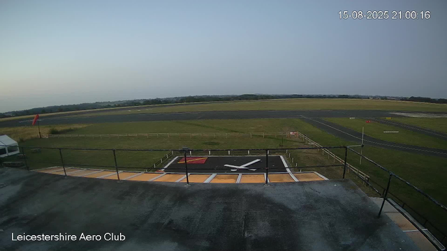 An empty helicopter landing pad with a large clock symbol painted on it, located on a rooftop. The horizon shows an open sky with some distant trees and fields, and the image has a timestamp of August 15, 2025, at 9:00 PM. The bottom left corner has the text "Leicestershire Aero Club."