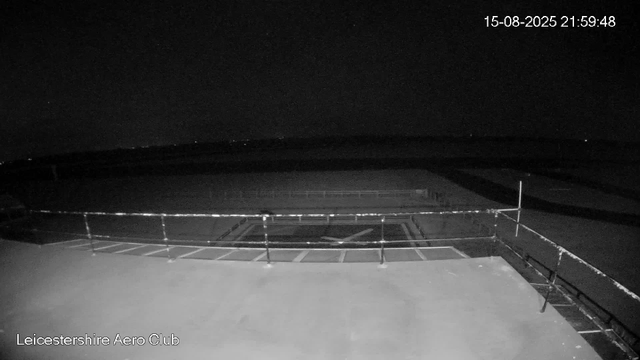 A black-and-white nighttime image from a webcam showing an empty outdoor area with a railing, likely a balcony or viewing platform at Leicestershie Aero Club. The scene is dark with no visible people or aircraft, and the sky appears clear with a few small stars visible. The date and time are displayed in the top right corner as 15-08-2025 21:59:48.