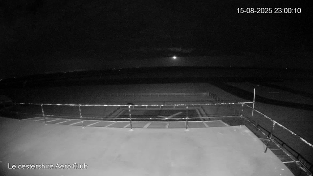 A night-time image from a webcam shows an empty outdoor area at Leicestershire Aero Club. The sky is dark with a bright moon partially obscured by clouds. In the foreground, there is a paved area with some metal fencing, and in the background, there are dark open fields or runways fading into the horizon. The timestamp in the top right corner reads "15-08-2025 23:00:10".