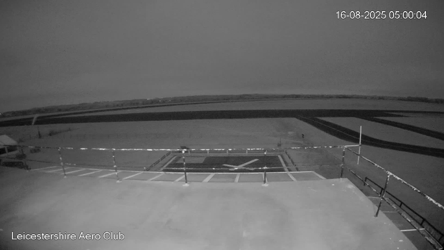 A black and white photo taken from a webcam at Leicestershire Aero Club at 5:00 AM on August 16, 2025. The image shows a small helicopter parked on a rooftop helipad with a white "X" marking. There are empty parking spaces with striped markings nearby, and a railing surrounds the helipad. Beyond the rooftop, open fields and a distant horizon are visible under a cloudy sky.