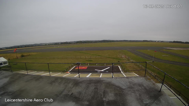 A view from a webcam at Leicestershire Aero Club showing an overcast sky above the runway and grassy area. In the foreground, there is a helipad with a large white "H" on a red background, surrounded by a black surface. The helipad is enclosed by a metal railing, and a small white tent is visible on the left side of the image. The image also includes a time stamp at the top right corner indicating the date and time as "16-08-2025 06:01:03". The overall scene appears calm with no aircraft or activity present.