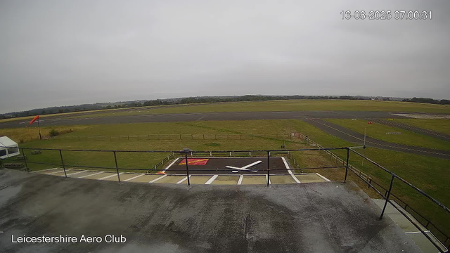 An empty helicopter landing pad with a red and white marking, surrounded by grass and a safety railing, overlooking an empty runway on a cloudy day at Leicestershire Aero Club. The timestamp shows the date and time as August 16, 2025, at 07:00:31.