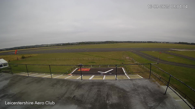 A view from a webcam at Leicester Aero Club shows an overcast sky above a runways and grass airfield area. In the foreground, there is a helipad marked with a large white "H" and a red square, surrounded by a fenced area. The tarmac on the rooftop where the camera is positioned is wet, indicating recent rain. The runway extends into the distance, with a windsock visible on the left side of the image and various runway markings visible on the pavement. The landscape beyond features open fields and some distant trees under a gray cloudy sky. The timestamp on the image indicates it was taken on August 16, 2025, at 8:01 in the morning.