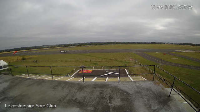 A view from a webcam at Leicestershire Aero Club shows the runway and surrounding area under overcast skies. In the foreground, there is a parking area with white lines and a helipad marked with a white "H" on a black surface, enclosed by a railing. A windsock on a pole is visible to the left, indicating wind direction. A small aircraft is parked on the runway in the background, and the landscape is mostly flat with grassy fields extending into the distance. The timestamp in the upper right corner reads "16-08-2025 10:00:38."