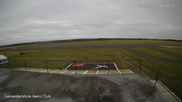 Overcast sky with a view of the runway and surrounding grassy areas at Leicester Airport. Visible tarmac and markings, including a helicopter landing pad in the foreground. Faint distant landscape in the background. The timestamp shows "16-08-2025 11:00:53".