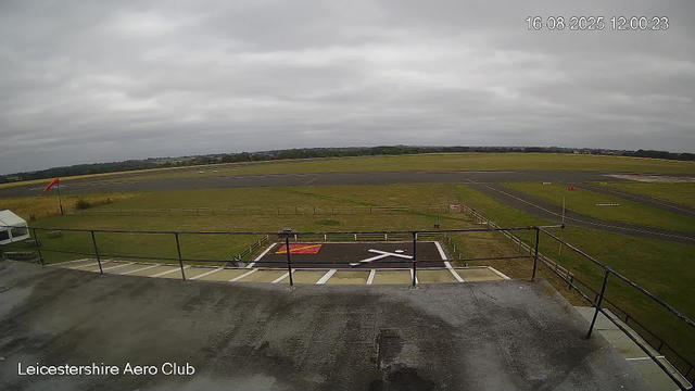 A view from a webcam at Leicestershire Aero Club shows an overcast sky with gray clouds. The airfield features a runway stretching across the horizon, surrounded by green grassy areas. In the foreground, there is a parking area with white painted lines and a designated helicopter landing zone marked with a large white "H" on a black background, enclosed by a metal railing. A windsock on a pole is visible on the left side of the image, indicating wind direction. The timestamp on the image reads 16-08-2025 at 12:00:23.