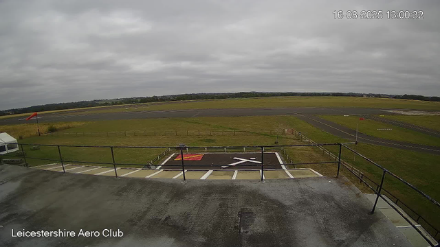 An overcast sky overlooking an airfield with a runway stretching across the background. In the lower part of the image, there is a helipad with a white and red markings, surrounded by a safety fence. To the left, a white tent is visible, and a windsock is positioned near the edge of the grassy area. The image is from the Leicesterhire Aero Club, and the timestamp shows it was taken on August 16, 2025, at 13:00.