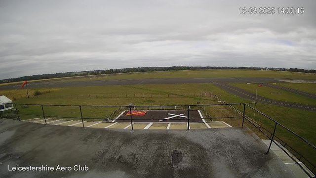 An outdoor view from a webcam at Leicestershire Aero Club on a cloudy day. The image shows the paved area and a helipad with a red and white symbol, adjacent to a grassy field. In the background, there is a runway with several markings, and the horizon features distant trees and a cloudy sky. The timestamp in the upper right corner indicates the date and time: August 16, 2025, at 14:00:16.
