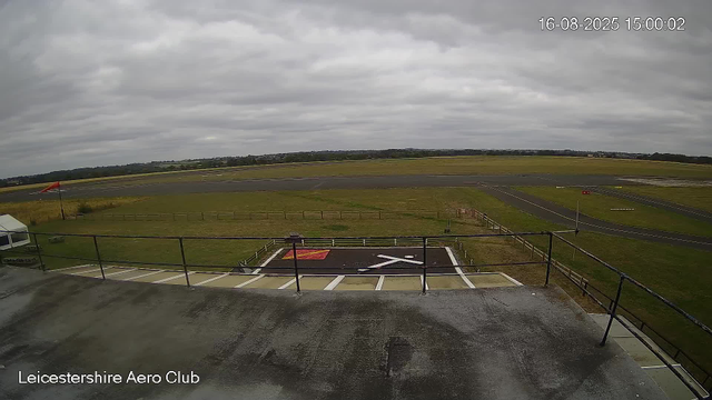 A view from a webcam at Leicesterhire Aero Club shows the tarmac runway and green grassy areas under a cloudy sky. There is a red windsock on the left, a black helipad with a white arrow and red markings in the foreground, and the surrounding open fields stretch out into the distance. The date and time in the upper right corner is 16-08-2025 at 15:00:02.