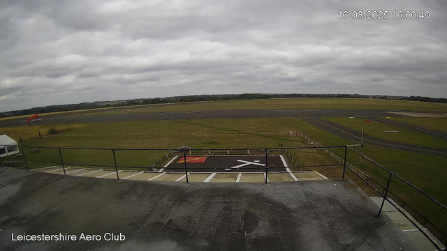 A view from a webcam at Leicesterhire Aero Club showing an overcast sky with gray clouds. The scene includes a runway and taxiways in the background, a fenced grassy area, and a helipad in the foreground with a red and black symbol. There is a small white tent on the left side of the image, and a windsock on a pole near the grass. The timestamp in the top right corner reads "16-08-2025 16:00:40."