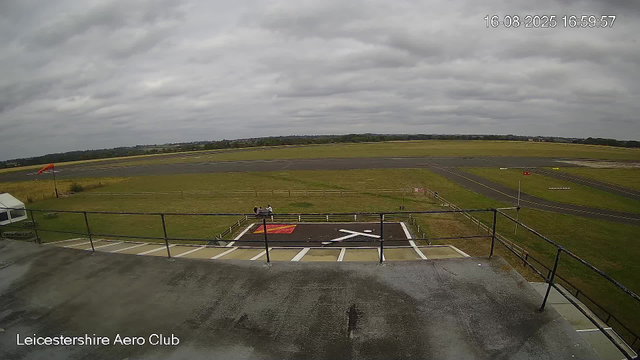 A view of an airfield taken from a webcam at Leicesterhire Aero Club. There is a grassy area, a small helipad with a white helicopter parked on it, and a paved runway stretching into the distance. The sky is overcast with gray clouds. In the foreground, there is a concrete platform with a black railing, and a person is seen near the helicopter. The timestamp shows the date and time as 16-08-2025, 16:59:57.