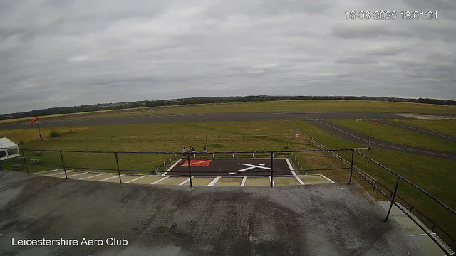 A view from a webcam at Leicesterhire Aero Club shows an overcast sky with gray clouds. In the foreground, there is a small helipad marked with a white helicopter symbol and a red and yellow square. Two people are sitting on a bench near the helipad. The runway and taxiway extend into the distance, with green grass surrounding the paved surfaces. There is a windsock on the left side of the image, and some fencing runs along the perimeter of the airfield. The image includes a timestamp showing "16-08-2025 18:01:01" in the upper right corner.