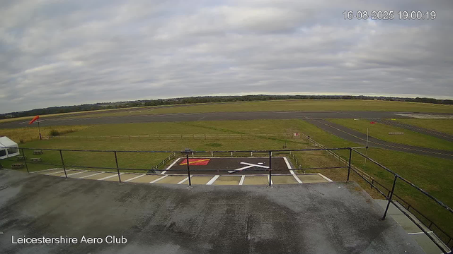 A view from a webcam at Leicestershire Aero Club shows the tarmac and runway area outdoors, with a grassy field extending into the distance under a cloudy sky. In the foreground, there is a helipad with a red and white marking, surrounded by a safety fence, and part of a small building or platform. The image is timestamped "16-08-2025 19:00:19".