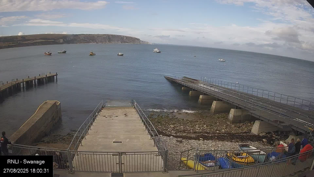 A coastal scene showing a small beach area with metal railings and concrete pilings, leading into calm water. Several small boats, including kayaks and dinghies, are stored on the shore. In the distance, there are multiple boats anchored or sailing on the water, with a hilly landmass in the background. A partly cloudy sky is overhead, and a few people can be seen near the boats on the right side of the image. The scene appears to be from a webcam, with a timestamp reading "27/08/2025 18:00:31" at the bottom left corner.