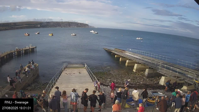 A group of people are gathered on a pier and rocky shoreline at a harbor, with some sitting on the edge and others standing or walking. Several boats are floating on the calm water in the harbor, with a distant coastline and hills visible under a partly cloudy sky. The scene appears lively with many visitors enjoying the area.