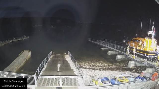 A small yellow rescue boat with several crew members on board is docked at a pier at night. The boat has tall antennas, safety lights, and a flag. The pier extends into the water, with a group of people in safety gear standing on it. There are boats and a fence visible in the foreground, and the scene is illuminated by artificial light, with a dark sky in the background. The timestamp indicates the image was taken on August 27, 2025, at 21:00:31.