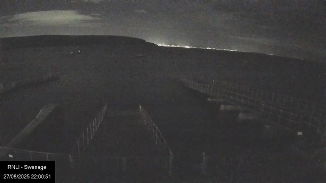 A dark, nighttime scene of a bridge or railway tracks seen from a webcam. The sky is cloudy with some faint light near the horizon. The image includes the text "RNLI - Swannage" and a timestamp "27/08/2025 22:00:51" in the bottom left corner.